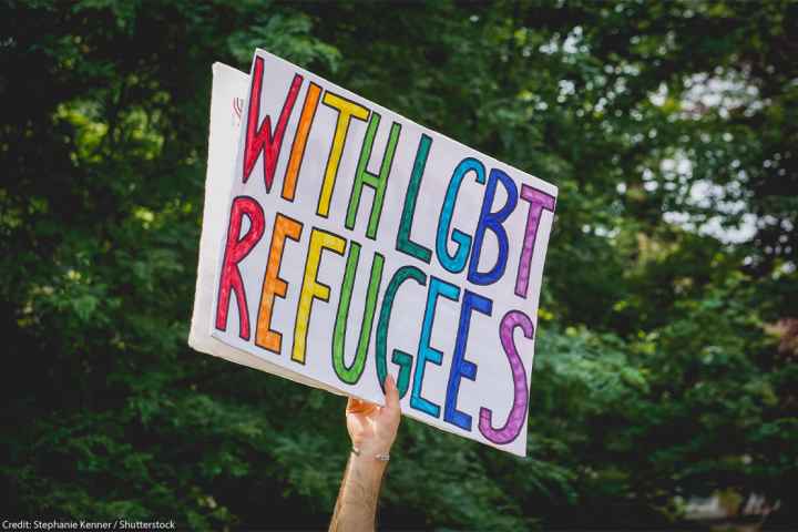 A hand holding up a "With LGBT Refugees" sign.