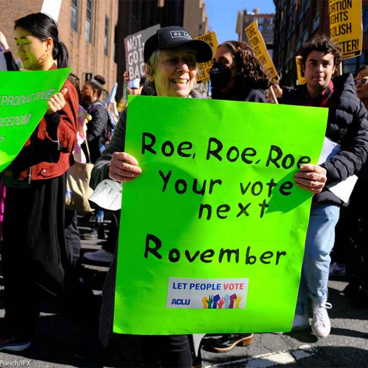 A woman holding a sign reading "Roe, Roe, Roe, Your vote next Rovember / LET PEOPLE VOTE ACLU" poses for the camera as fellow activists walk behind her.