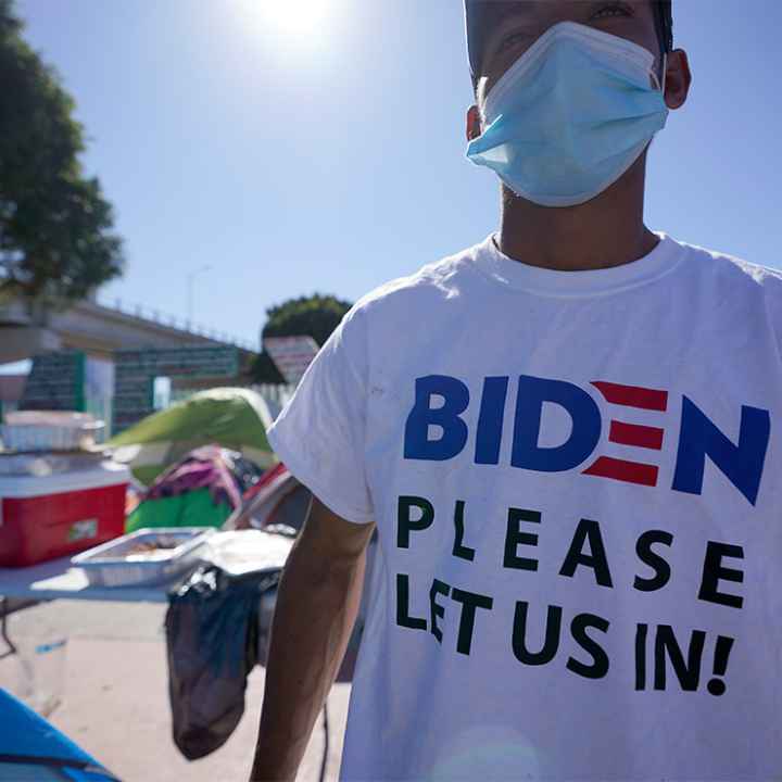 A man seeking asylum in the United States wears a shirt that reads, "BIDEN PLEASE LET US IN!," as he stands among tents that line an entrance to the border crossing in Tijuana, Mexico.