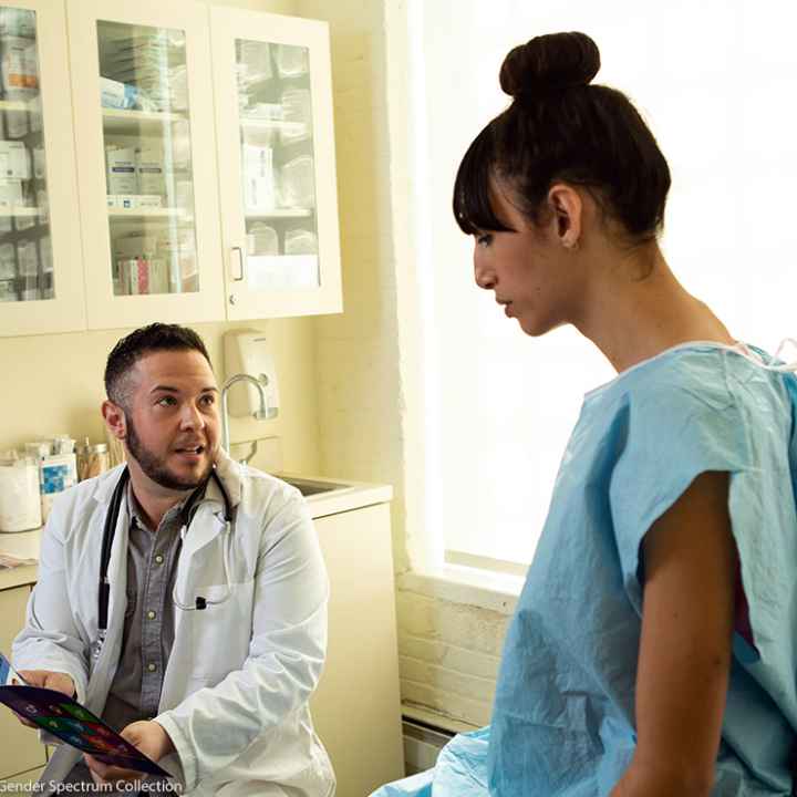 A transgender woman in a hospital gown speaking to her doctor, a transgender man, in an exam room