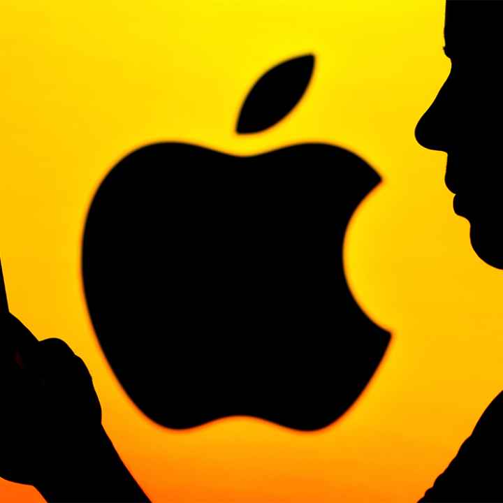 In shadow, a person holds a phone with Apple logo (also in shadow) against a yellow background.