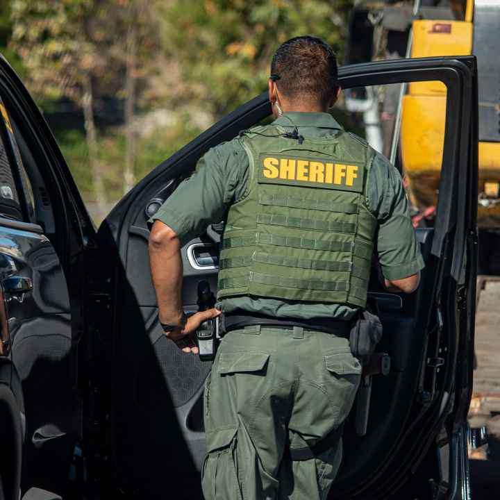 Shot of sheriff (wearing a bulletproof vest) from behind walking towards the open door of police car.