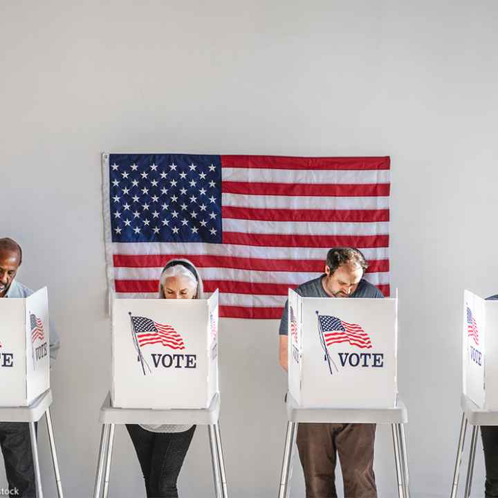 With and American flag in the background, four people in the act of voting, stand behind voter booths as they make their selections.